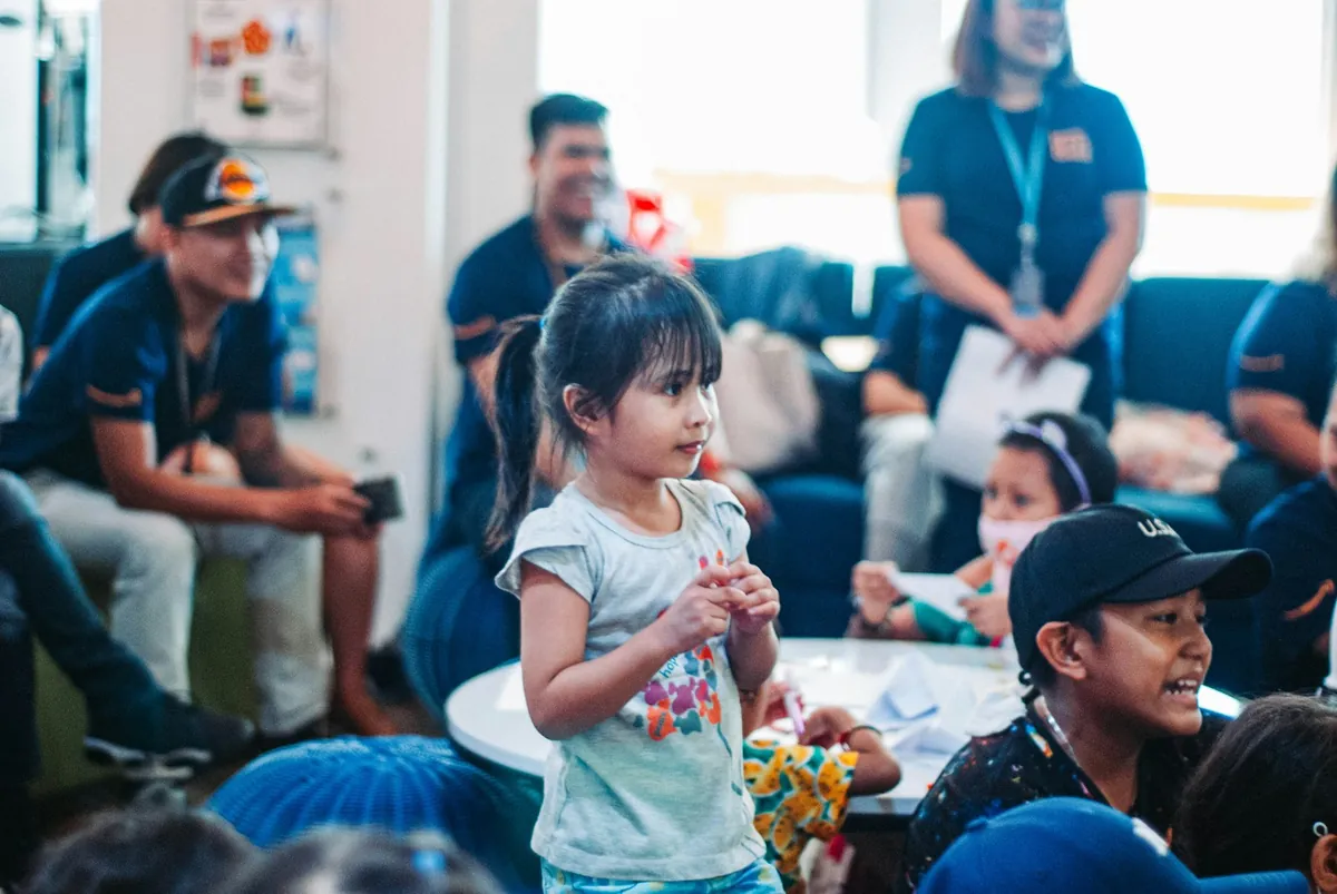 A child standing at a party with adults sitting nearby