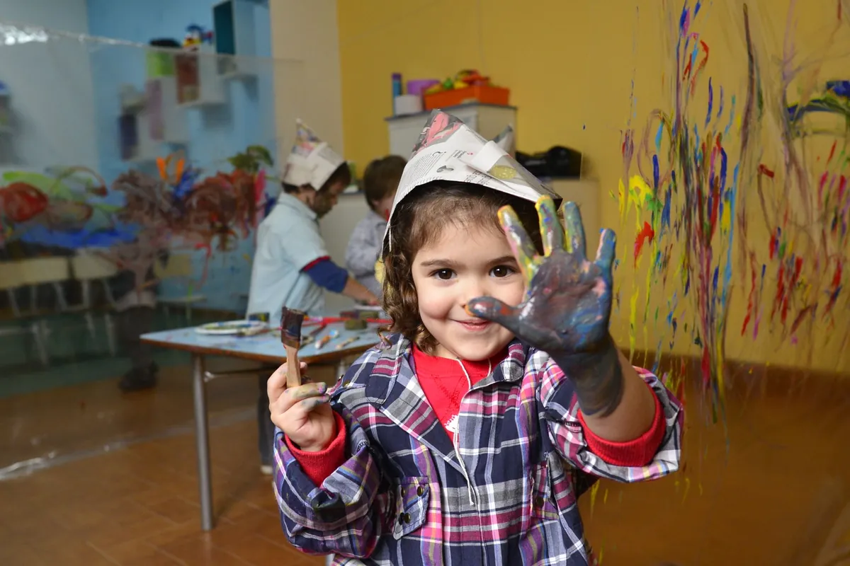 A young child holding up paint-covered hands in a playroom