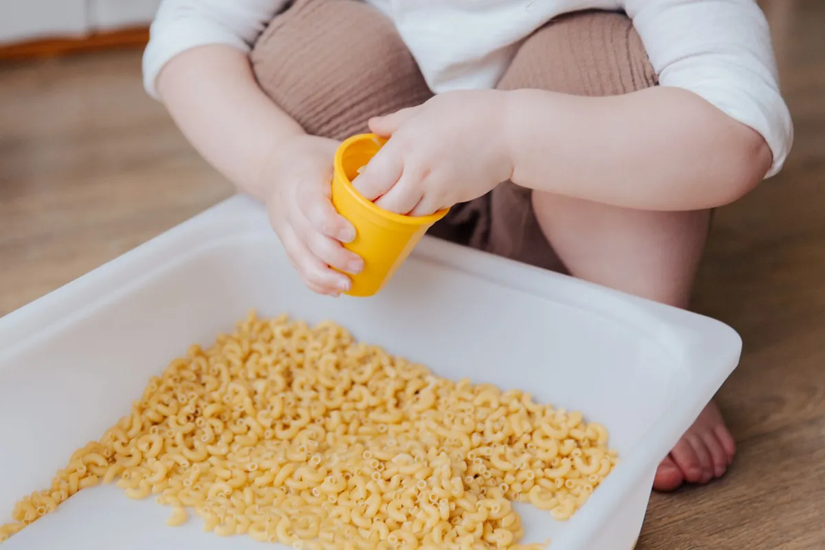 Small hands scooping dried pasta from a shallow tray