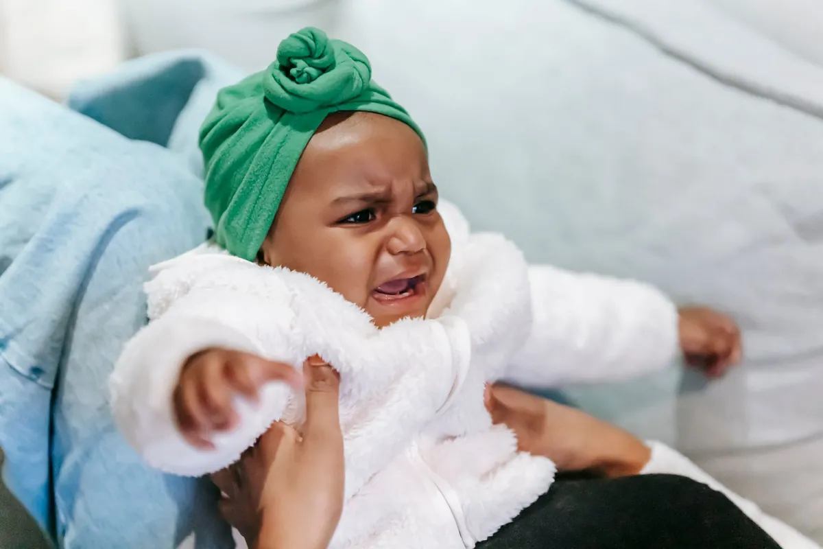 An upset baby in a green hat held in a parent arms