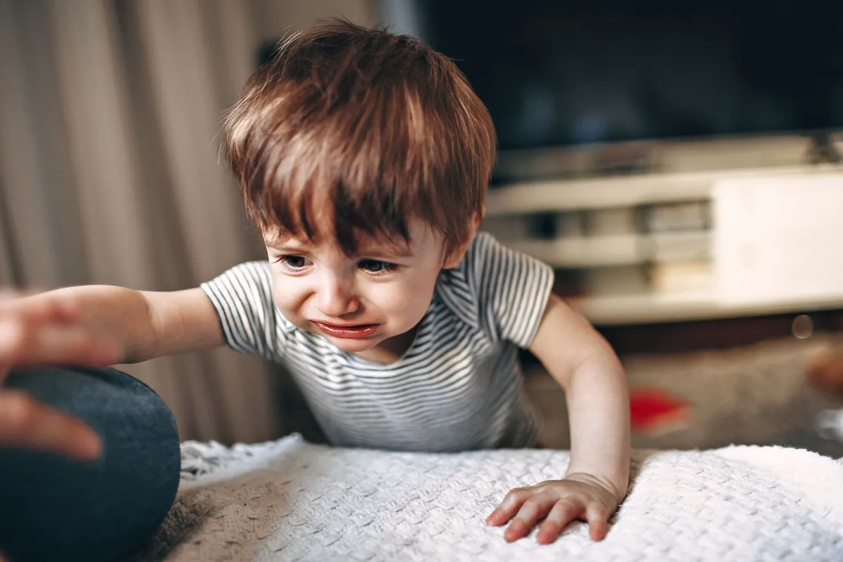 An upset toddler reaching out while sitting on a blanket