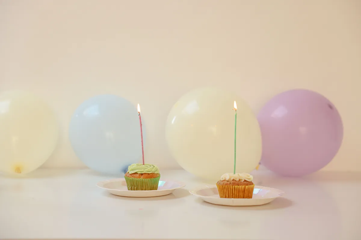Two cupcakes with lit candles in front of pastel balloons