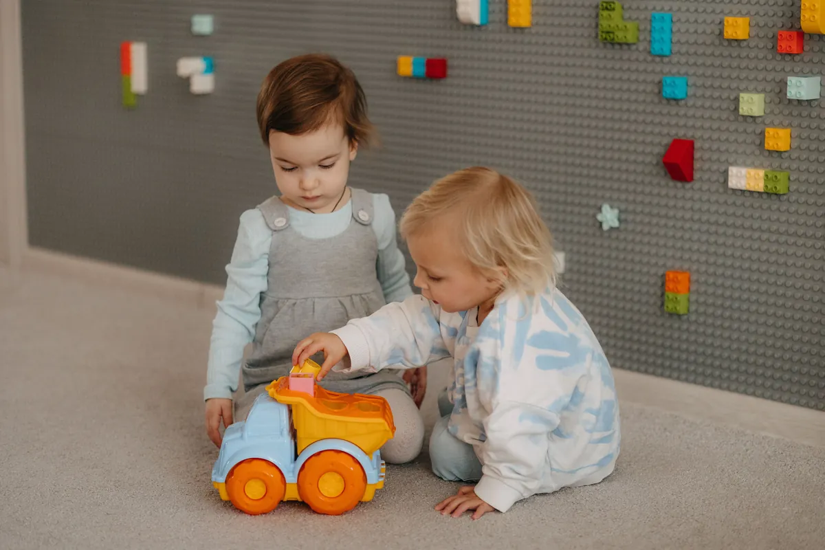 Two toddlers playing side by side with toy cars on the floor